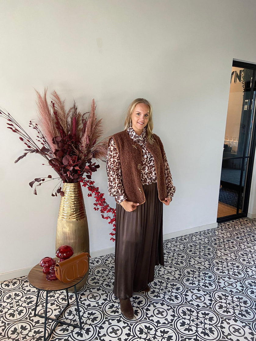 Een vrouw staat binnen op tegels met een patroon en draagt een vest van bruin imitatiebont, een blouse met luipaardprint en de Bruine rok met elastische tailleband, gecombineerd met bruine laarzen. Naast haar staan een gouden vaas met gedroogde bloemen en een tafel met een bruine handtas.