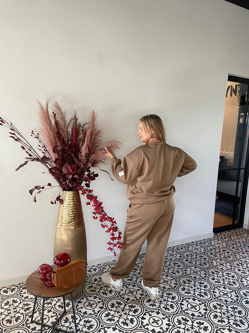 Een vrouw in Outfit - bruin kijkt met haar rug naar de camera naar een beige muur, naast een hoge gouden vaas met rode droogbloemen op tegels met een zwart-wit patroon. Een kleine tafel met rood decor staat vlakbij.