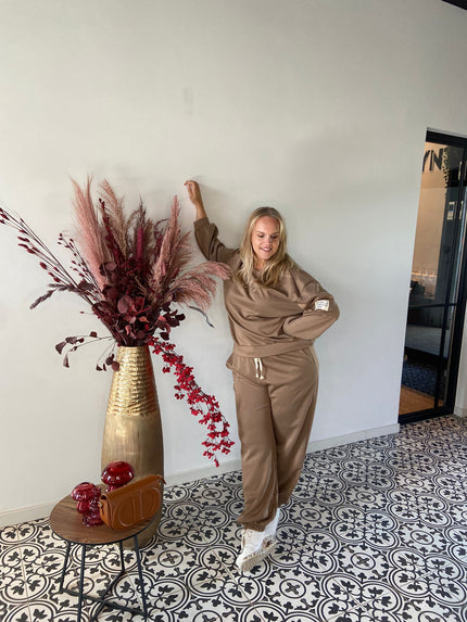 Een vrouw in de Outfit - bruin trainingspak en witte sneakers staat bij een grote gouden vaas met bordeauxrode en roze droogbloemen, bij een tafeltje met een rode handtas, op een tegelvloer met patroon tegen een effen lichte muur.