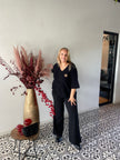 Een blonde vrouw staat binnen naast een gouden vaas met gedroogde bloemen op een tafeltje, ze draagt de Trui - Zwart van Bourgondië met driekwart mouw. De vloer heeft zwarte en witte tegels. One size past 38-48, ideaal voor herfst winter 2025.