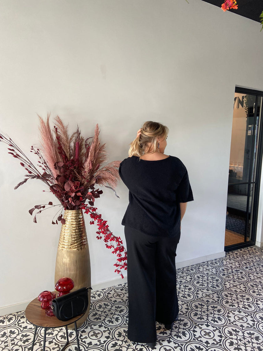 Een vrouw met blond haar staat met haar rug naar de camera en draagt een Trui - Zwart van Bourgondië (een maat 38-48), gecombineerd met een zwarte broek. Naast haar staat een gouden vaas met gedroogde bloemen en een tafeltje met rode ornamenten en een zwarte tas. Herfst winter 2025.