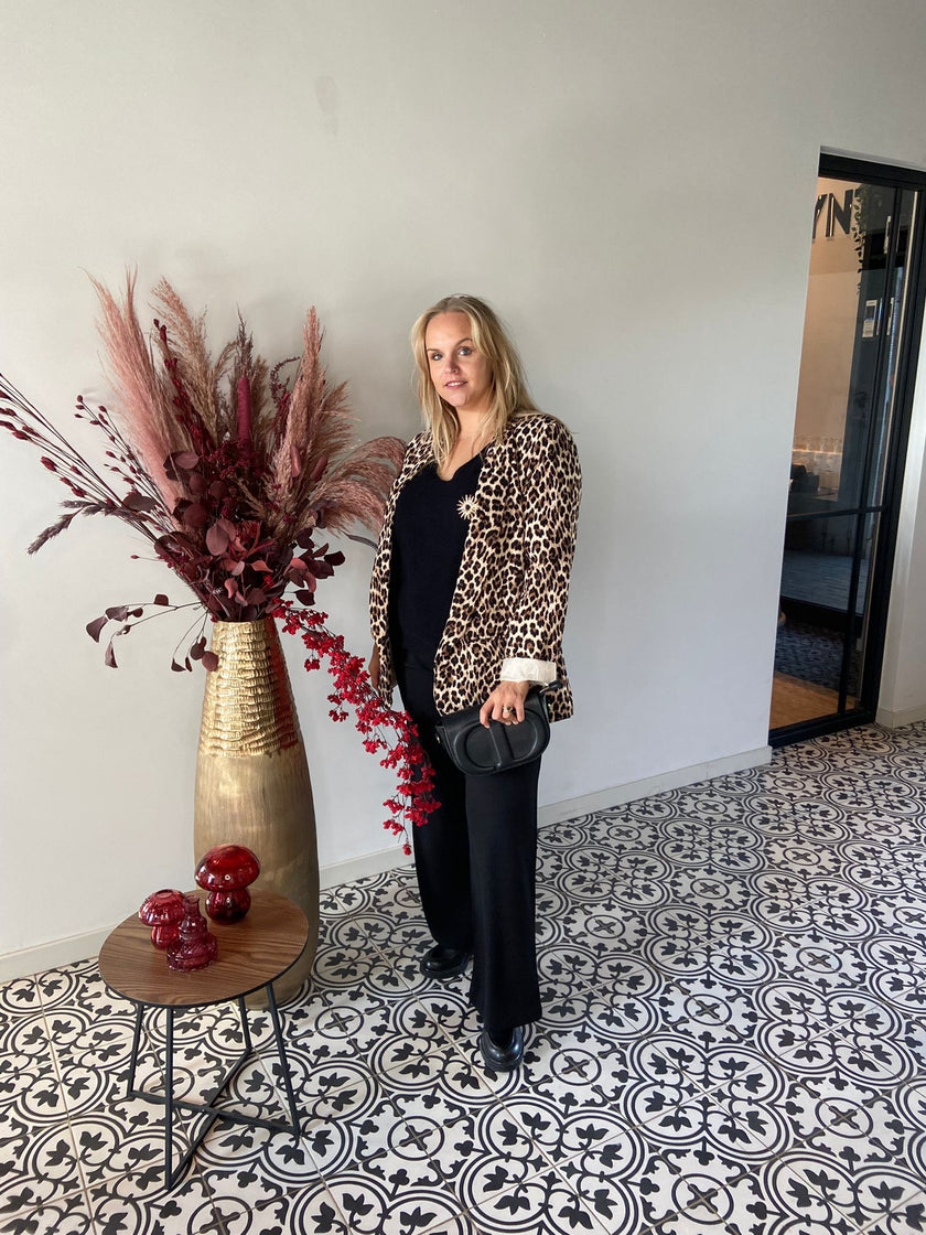 Een vrouw met de Blazer met print en een zwarte broek staat binnen op een tegelvloer met patroon en houdt een zwarte tas vast. Naast haar staan een hoge vaas met gedroogde planten en een kleine ronde tafel met rode ornamenten.