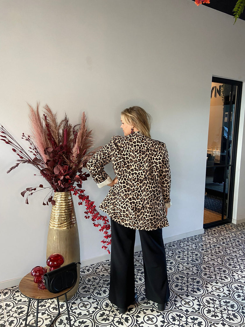 Een vrouw staat met haar rug naar de camera, draagt de Blazer met print en een zwarte broek naast een hoge gouden vaas met bordeauxrode en roze droogbloemen, op een tegelvloer met patroon bij een tafeltje met een zwarte tas.
