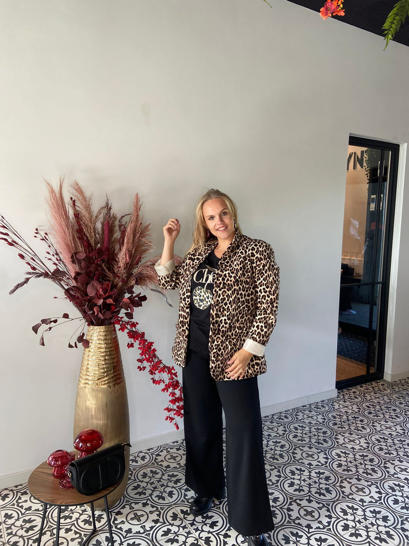 Een vrouw staat binnen op tegels met een patroon en draagt een blazer met luipaardprint, een zwarte broek met wijde pijpen en het V-hals T-shirt kersenzwart. Ze poseert bij een hoge gouden vaas met gedroogde rode en roze bladeren - een elegante look voor herfst/winter 2025.