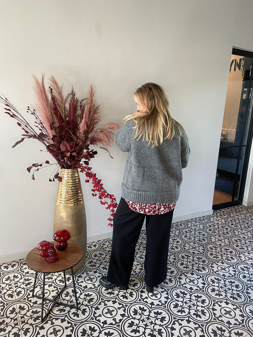 Een blonde vrouw in een grijze grofgebreide trui en zwarte broek staat binnen bij de Olieve Essential-een hoge gouden vaas met rode en roze gedroogde bloemen-bij een tafeltje met rode ornamenten op een tegelvloer met patroon.