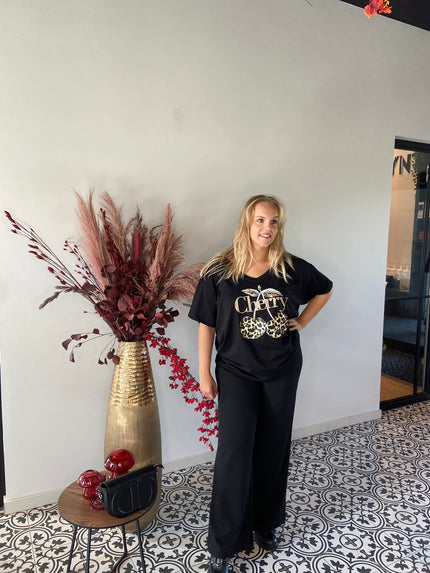 Een blonde vrouw staat binnen op gedessineerde tegels, draagt het plus-size V-hals T-shirt kersenzwart en een zwarte broek. Ze poseert naast een hoge gouden vaas met bordeauxrode gedroogde planten en een tafel met een zwarte tas en rood decor.