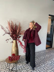 Een vrouw draagt de jas Beatrice Jas - Bordeaux en een zwarte broek. Ze staat binnen op tegels met een patroon naast een hoge gouden vaas met bordeauxrode droogbloemen en een tafeltje met bijpassend decor.