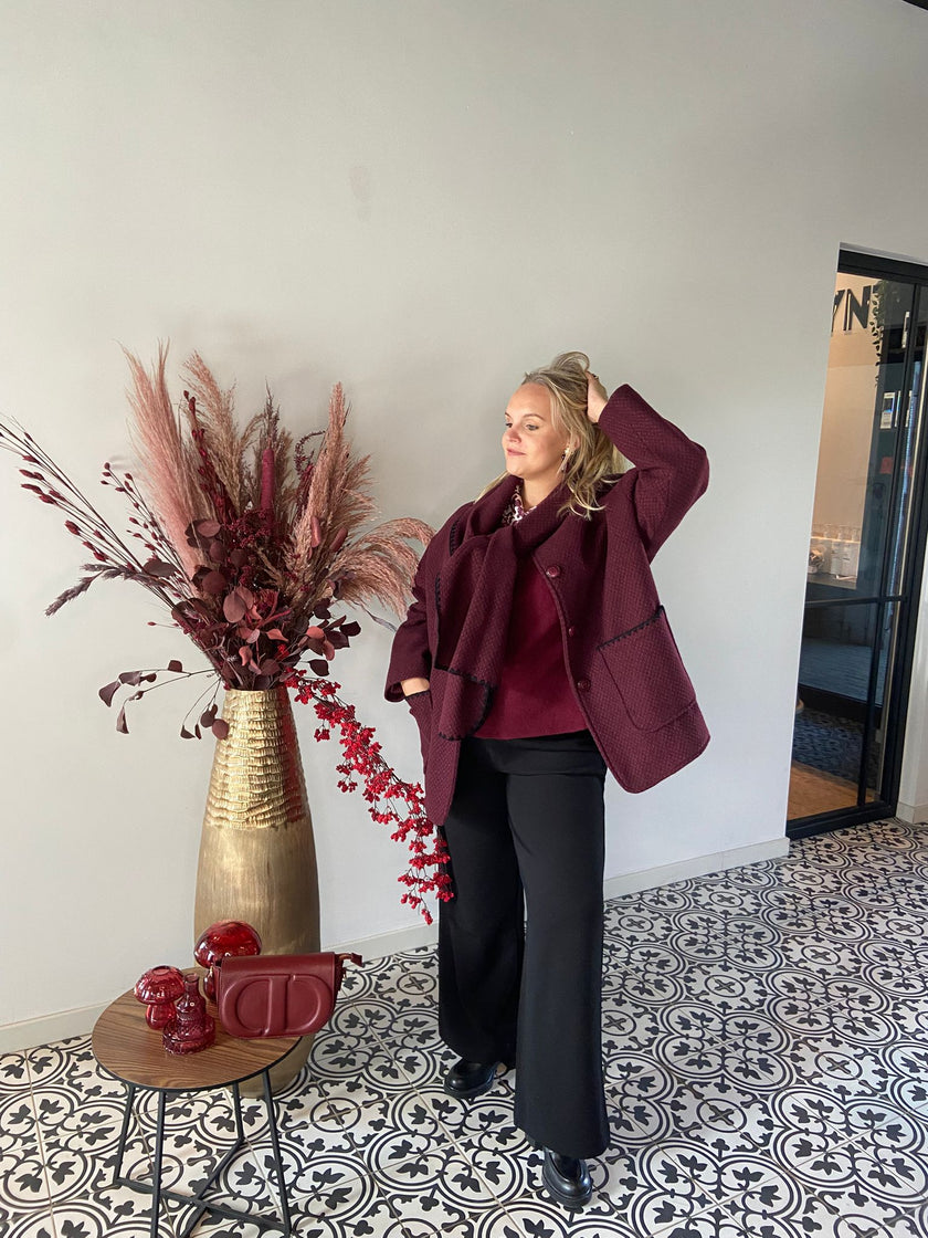Een vrouw draagt de jas Beatrice Jas - Bordeaux en een zwarte broek. Ze staat binnen op tegels met een patroon naast een hoge gouden vaas met bordeauxrode droogbloemen en een tafeltje met bijpassend decor.