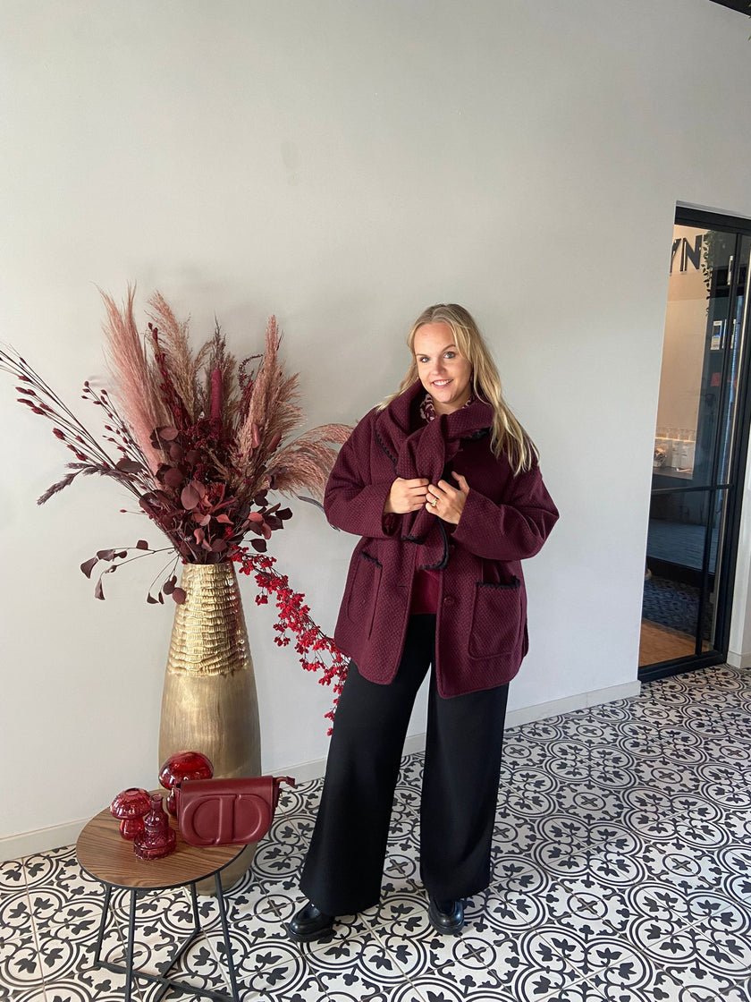 Een vrouw met de Beatrice Jas - Bordeaux staat binnen op een tegelvloer met patroon naast een gouden vaas met gedroogde bloemen en een kleine ronde tafel met rode decoratieve stukken.
