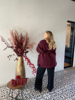Een vrouw met lang blond haar draagt de Beatrice Jas - Bordeaux en een zwarte broek. Ze staat binnen bij een grote gouden vaas gevuld met rode en roze droogbloemen, naast een tafeltje met rood decor op vloertegels met patroon.
