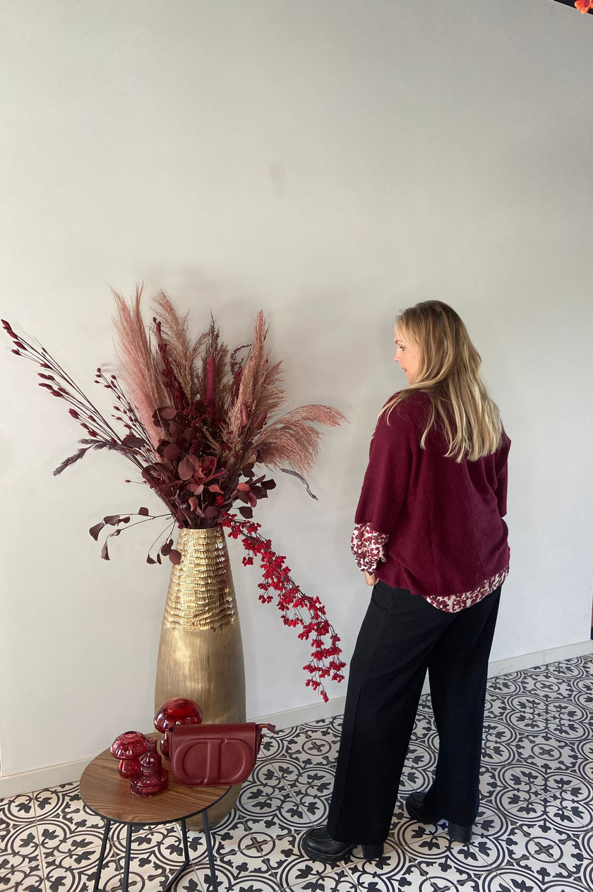Een vrouw met lang blond haar staat binnen voor een hoge gouden vaas met kastanjebruine en roze droogbloemen. Ze draagt de Trui - Zwart van Bourgondië, een maat 38-48 met een gebloemde zoom, een zwarte broek en een kastanjebruine tas op een tafeltje.