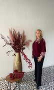 Een vrouw met lang blond haar modelleert de Trui - Zwart of Burgundy (een maat 38-48, driekwart mouw), gecombineerd met een zwarte broek, staand binnen naast een hoge gouden vaas met bordeauxrode en roze droogbloemen-ideaal voor herfst winter 2025.