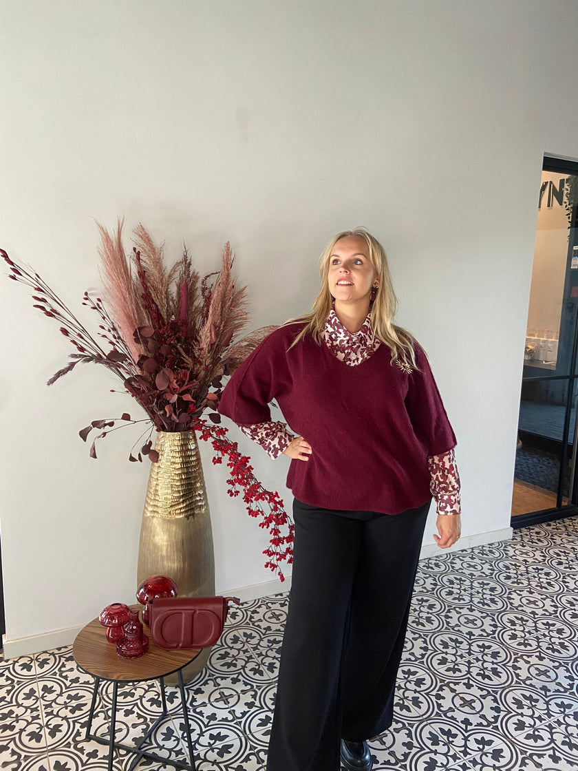 Een vrouw staat binnen op een tegel met patroon en draagt de Trui - Zwart of Burgundy over een blouse met patroon en een zwarte broek. Ze poseert bij een hoge vaas met gedroogd rood gebladerte, wat deze gezellige herfstlook van 2025 benadrukt. Eén maat: 38-48.