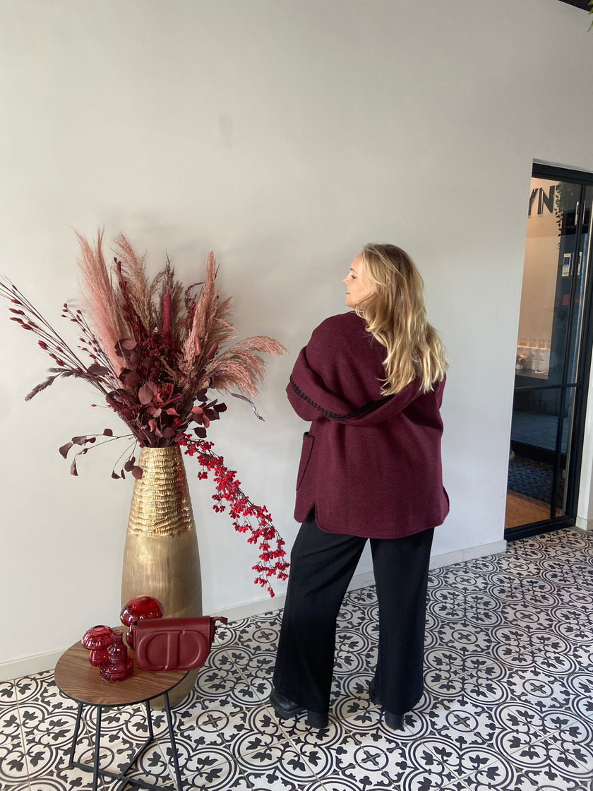 Een vrouw met lang blond haar draagt de Beatrice Jas - Bordeaux en een zwarte broek. Ze staat binnen bij een grote gouden vaas gevuld met rode en roze droogbloemen, naast een tafeltje met rood decor op vloertegels met patroon.