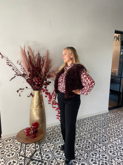Een vrouw met lang blond haar staat op tegels met een patroon en draagt het Caroline - Dark Bordeaux vestje over een bijpassende blouse en zwarte broek. Ze poseert bij een gouden vaas met gedroogd bordeauxrood en roze gebladerte, haar look stijlvol gelaagd.