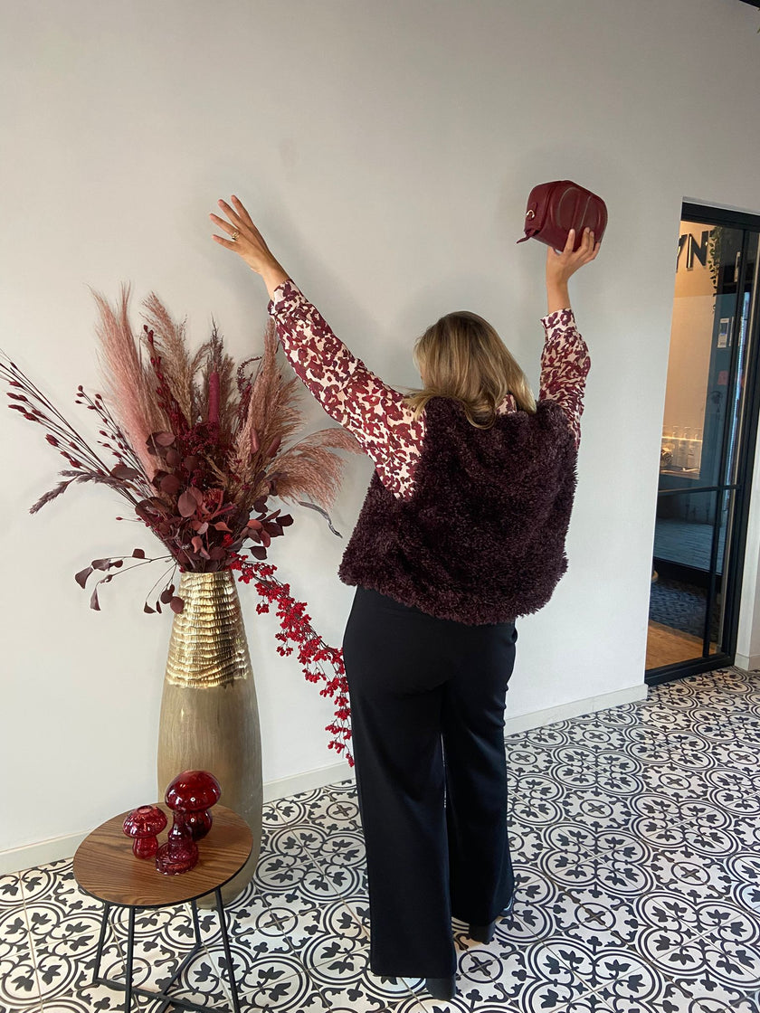 Een vrouw met haar rug naar de camera draagt de Caroline - Dark Bordeaux, een kleine tas, en staat in zwarte broek, vestje en een kort, zacht, gepatroneerd bloesje naast een hoge vaas met rode en roze gedroogde bloemen op patroontegels.