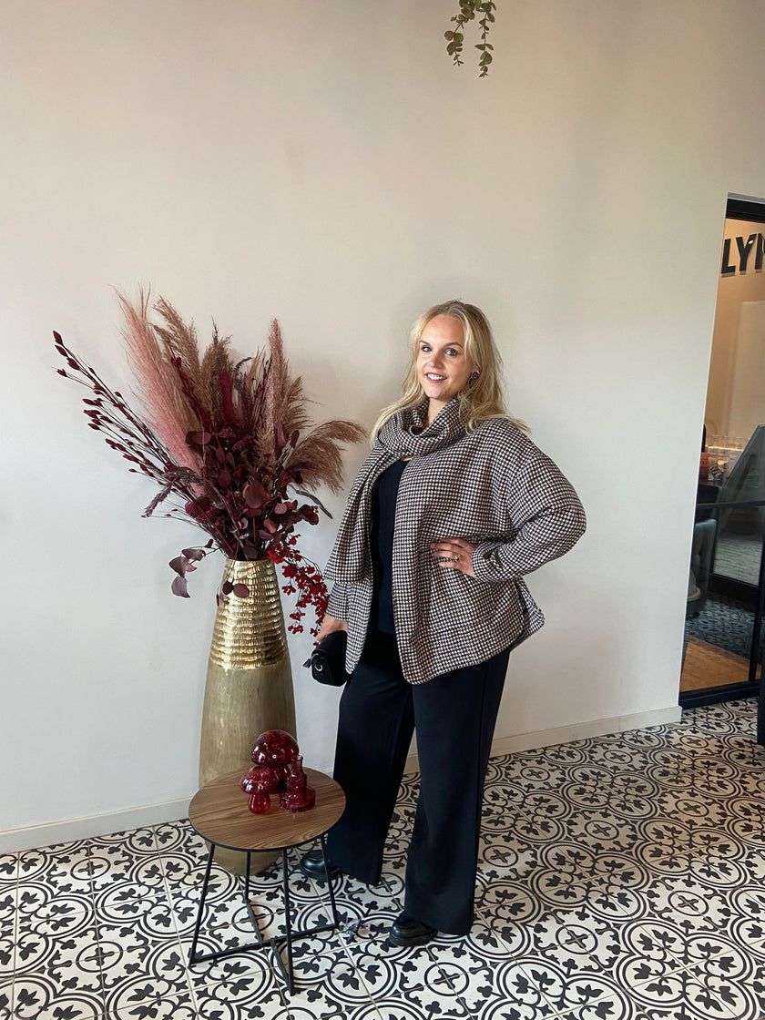 Een vrouw met lang blond haar staat binnen op een decoratieve tegelvloer, poserend in de Adele Jas - Amagia houndstooth jas en zwarte outfit naast een hoge gouden vaas gevuld met rode en roze gedroogde bladeren op een kleine ronde tafel.