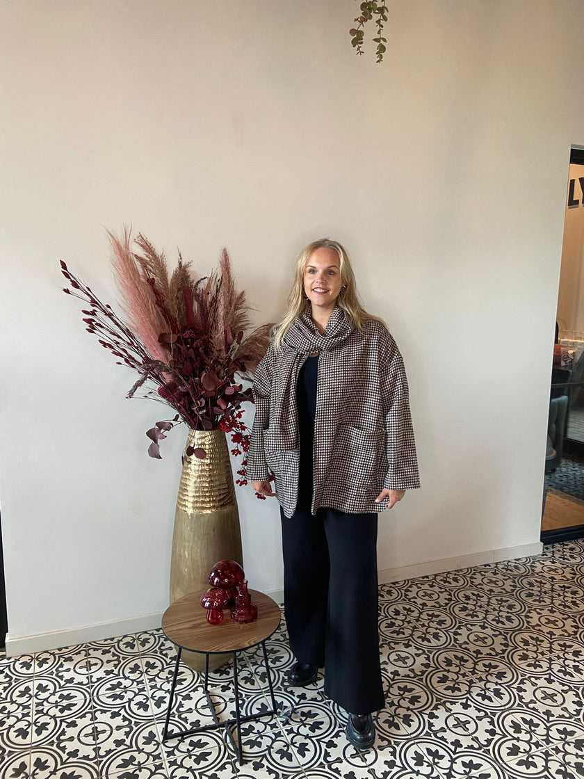 Een vrouw met lang blond haar draagt de geruite jas en zwarte broek van Adele Jas - Amagia en glimlacht binnenshuis naast een hoge gouden vaas met gedroogde bordeauxrode planten en een tafeltje met rood decor op vloertegels met patroon.