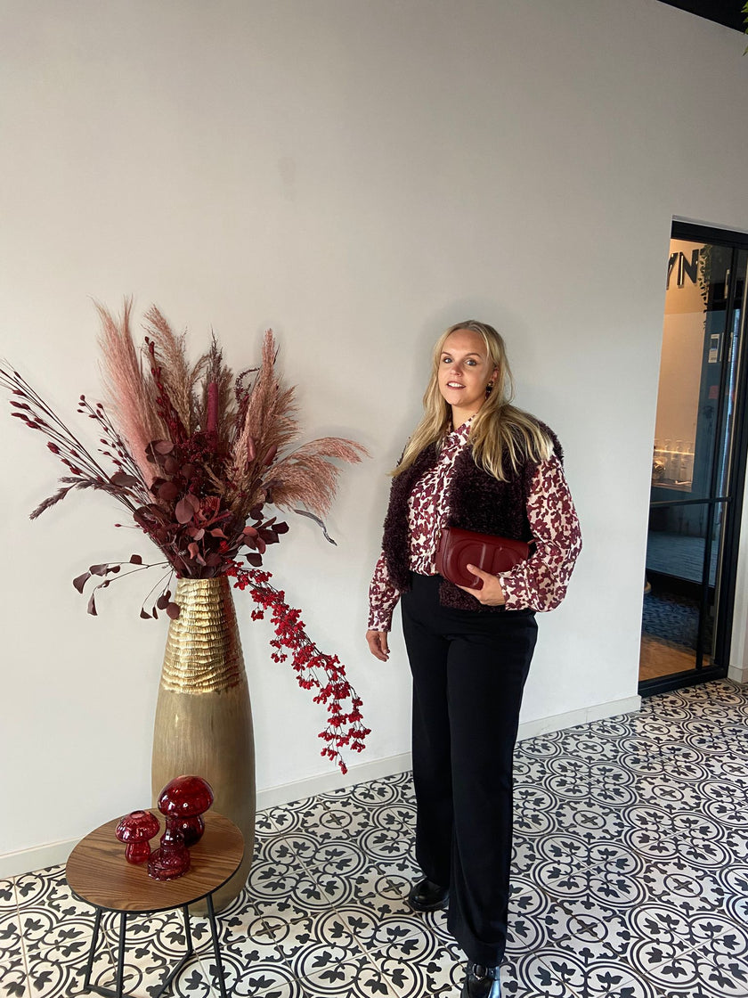 Een vrouw met lang blond haar staat binnen bij een hoge vaas met gedroogde bloemen. Ze styled haar look met een blouse met patroon, een zwarte broek en de Caroline - Dark Bordeaux clutch op een zwart-witte tegelvloer.