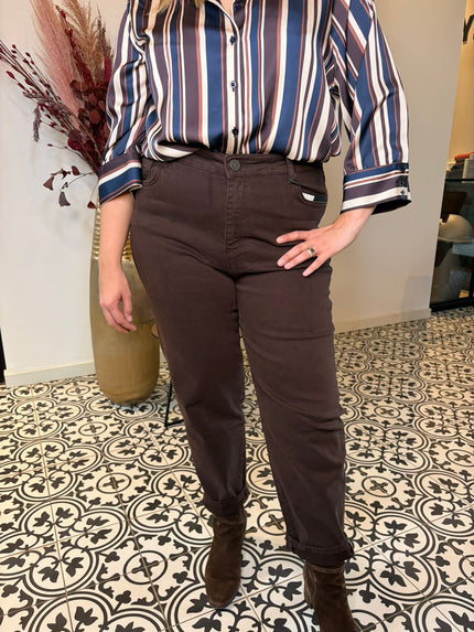 Een persoon staat binnen in een gestreept shirt, Bruine jeansbroek - Mat Fashion en bruine laarzen. Een hand rust op de heup, met gedessineerde vloertegels en een hoge vaas met gedroogde planten op de achtergrond, in een chique MAT Fashion-stijl.