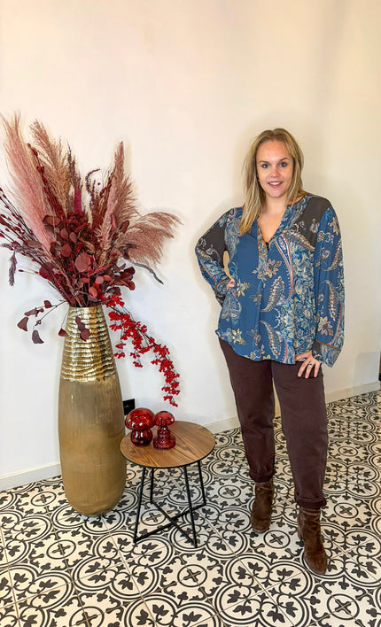 Een vrouw met blond haar draagt de Bohemian blouse van Mat Fashion, met een blauwe print en wijde mouwen, in combinatie met een bruine broek. Ze staat binnen op gedessineerde tegels naast een gouden vaas met gedroogd rood gebladerte en een tafeltje met rood decor.