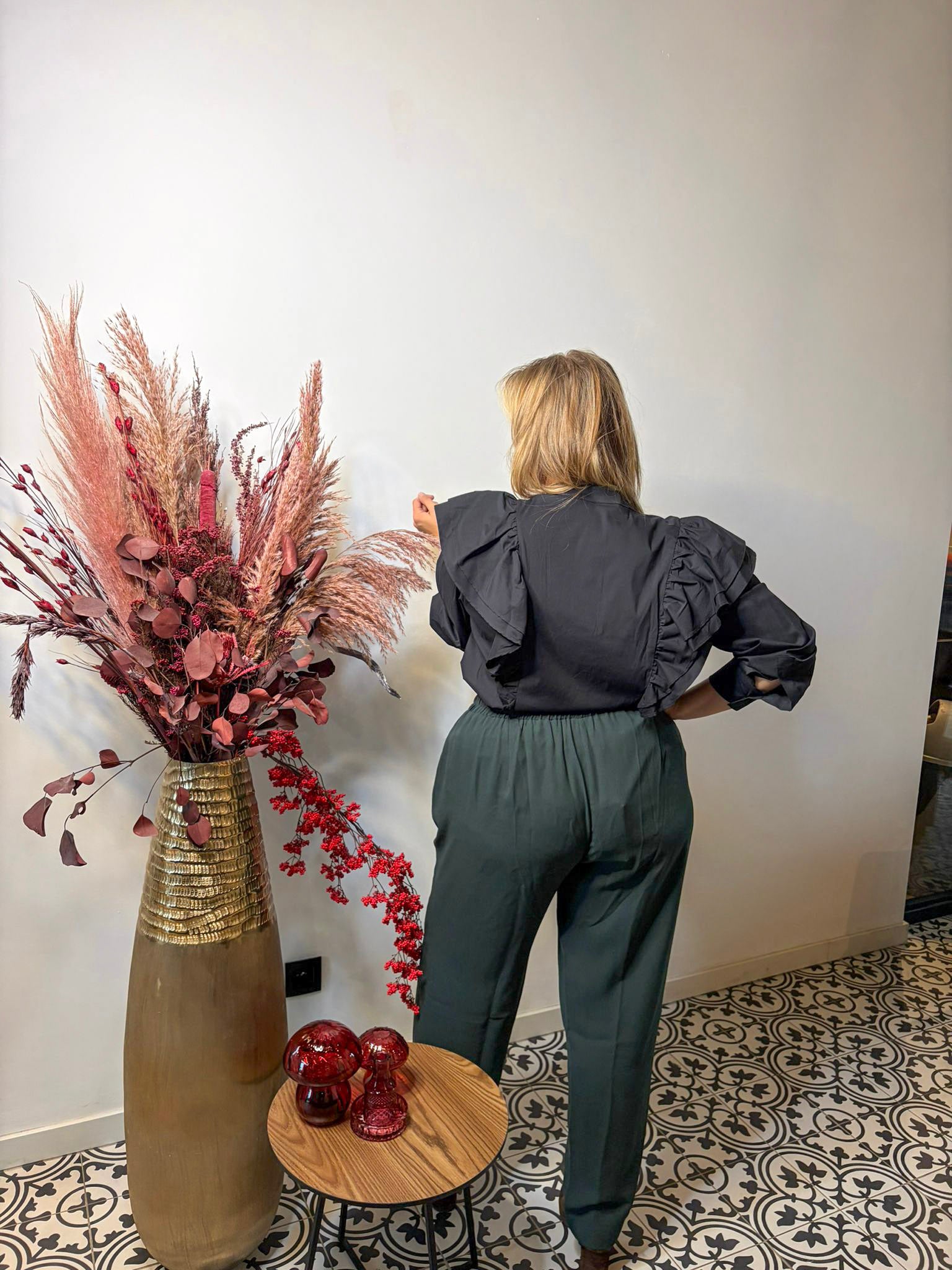 Een vrouw kijkt weg van de camera en draagt de Zwart hemd van Mat Fashion - een zwarte blouse met ruches en franjé-effect - in combinatie met een groene broek met hoge taille. Naast haar staat een tafel gedecoreerd met rode glazen stukken en een hoge vaas met gedroogde roze en rode bloemen.