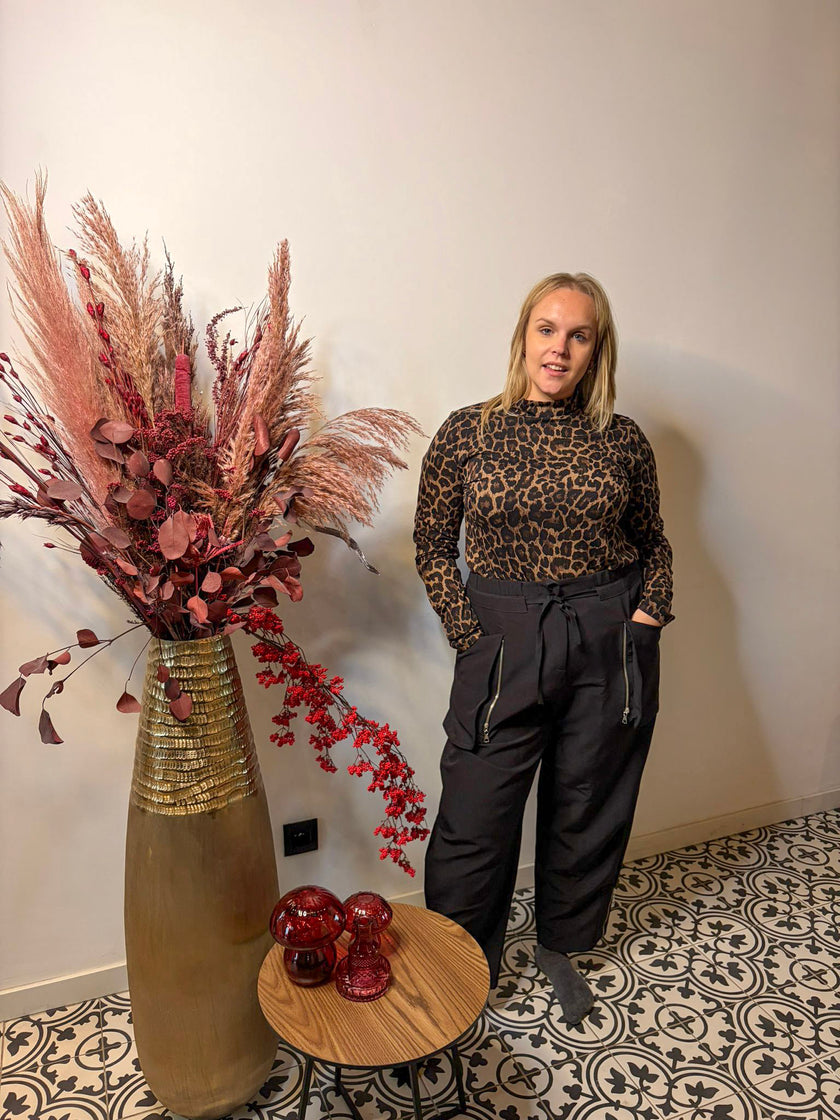 Een vrouw draagt de Zwarte Broek van Mat Fashion en een top met luipaardprint. Ze staat blootsvoets binnen, handen in de zakken, naast een grote vaas met gedroogde bloemen in een modern ingerichte kamer met een rond houten tafeltje.