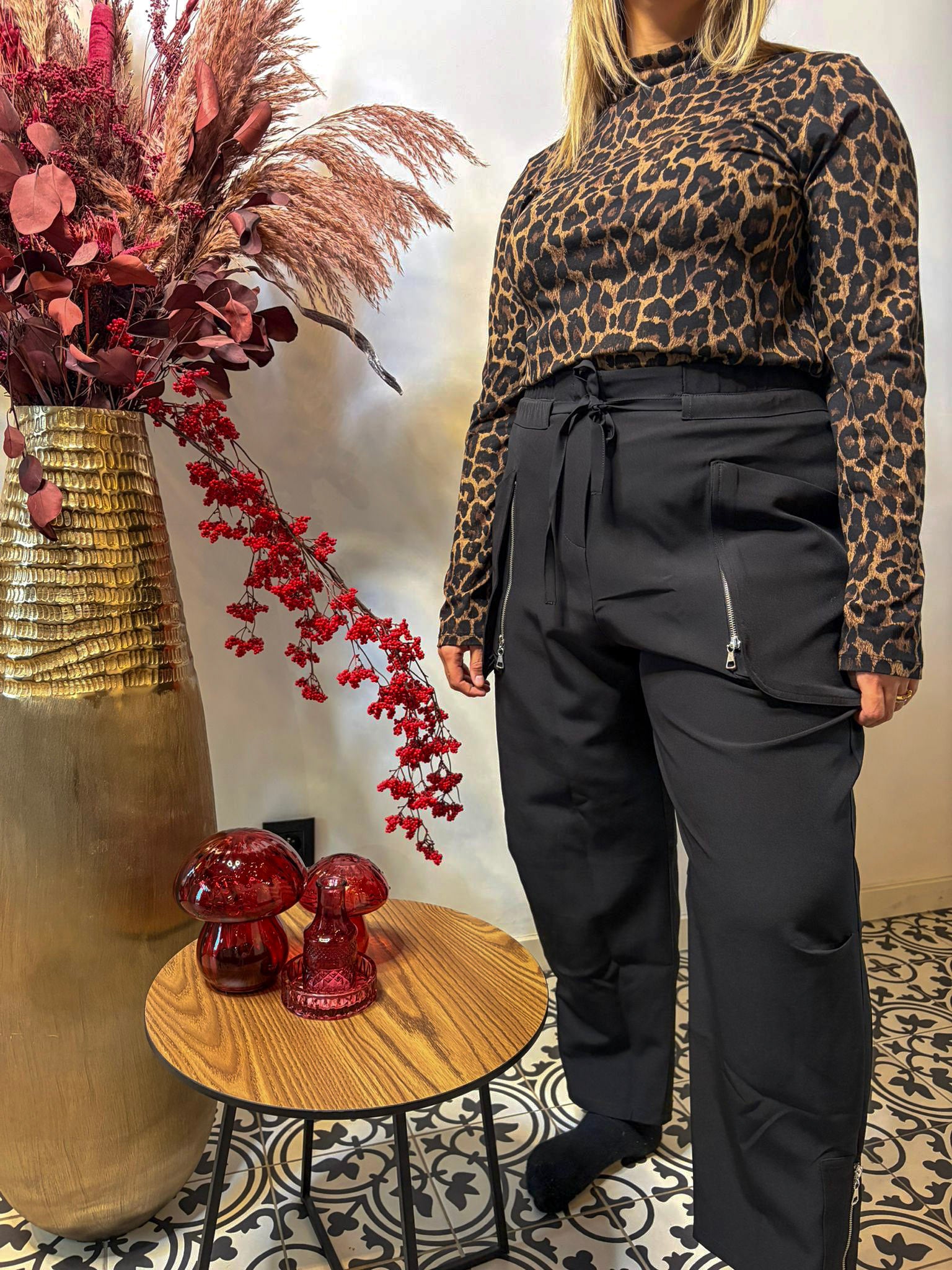 Een vrouw binnenshuis draagt een coltrui met luipaardprint en een Zwarte Broek - Mat Fashion, met een moderne uitstraling. Naast haar staan een gouden vaas met gedroogde bloemen en een tafeltje met rood glazen decor op gedessineerde tegels.