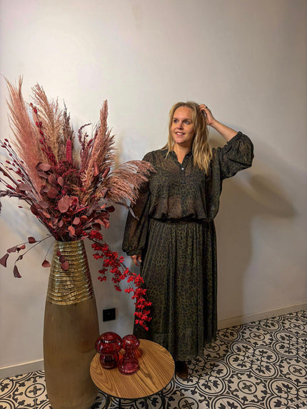 Een vrouw in de Donkergroene rok met print - Mat Fashion staat binnen naast een hoge vaas met pampagras en rood gebladerte, glimlacht en raakt haar haar aan bij een kleine ronde tafel met rood glazen decor op vloertegels met patroon.