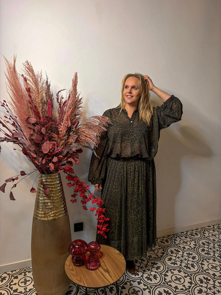 Een vrouw in de Donkergroene rok met print - Mat Fashion staat binnen naast een hoge vaas met pampagras en rood gebladerte, glimlacht en raakt haar haar aan bij een kleine ronde tafel met rood glazen decor op vloertegels met patroon.