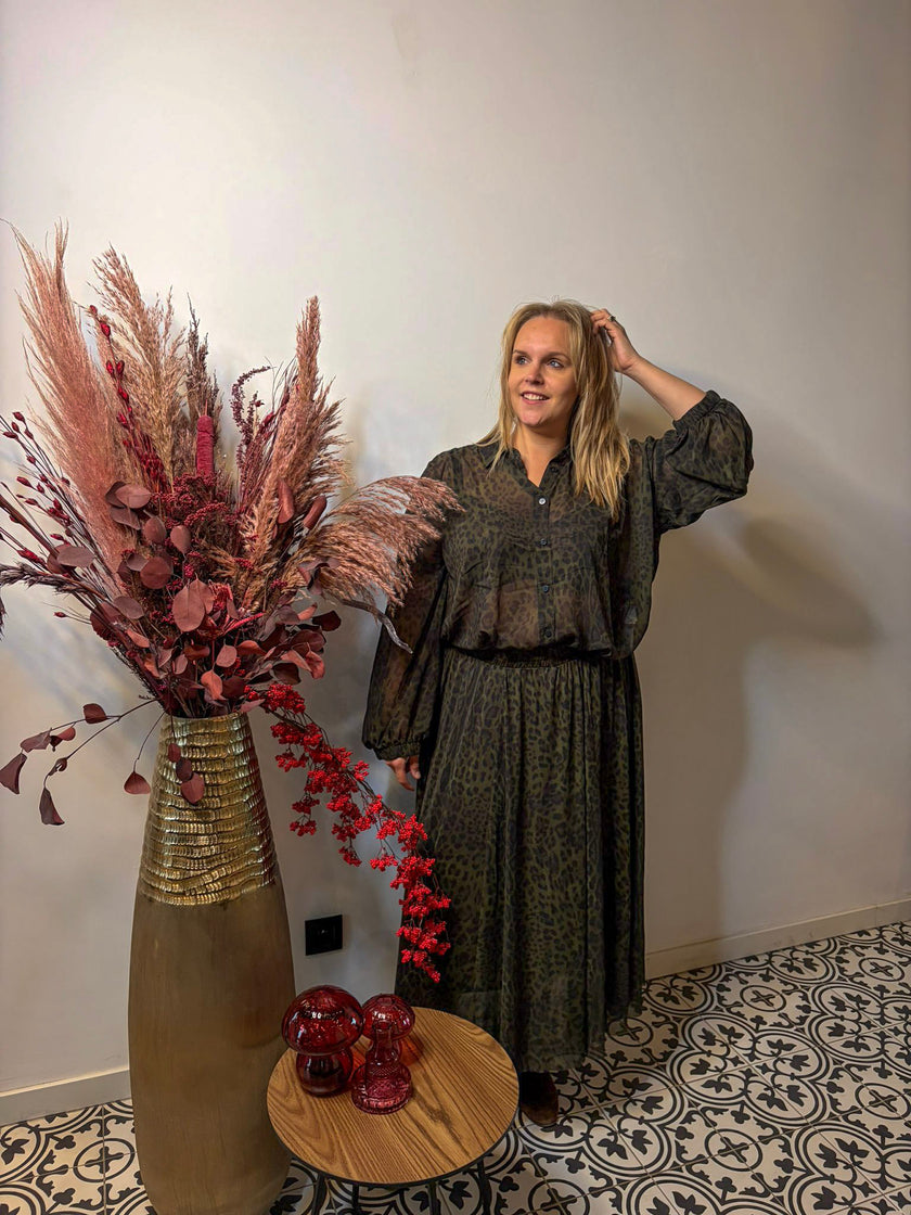 Een vrouw in de Donkergroene rok met print - Mat Fashion staat binnen naast een hoge vaas met pampagras en rood gebladerte, glimlacht en raakt haar haar aan bij een kleine ronde tafel met rood glazen decor op vloertegels met patroon.