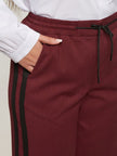 Iemand draagt een Rode broek met strepen - Mat Fashion, een stijlvolle kastanjebruine broek met zwarte strepen en een koord in de taille voor een moderne sportieve look, gecombineerd met een wit overhemd en één hand in de zak.