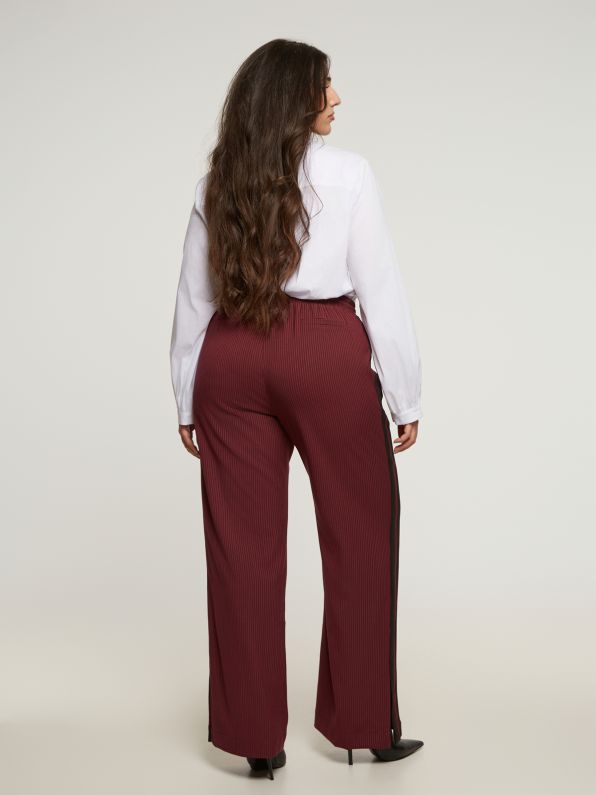 Vrouw met lang bruin haar draagt een witte blouse en Rode broek met strepen - Mat Mode, staand en met het gezicht van de camera weg op een effen lichte achtergrond.