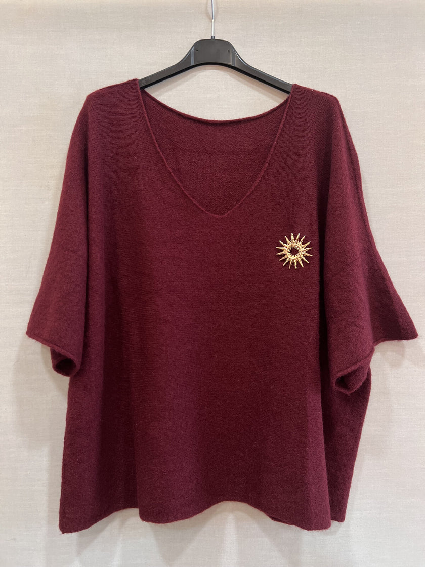 De Trui - Zwart of Burgundy is een ruimvallende, kastanjebruine top met korte mouwen, een V-hals en een gouden zonnestraalbroche op de linkerborst. Perfect voor de winter van 2025, biedt dit stuk in maat 38-48 moeiteloos comfort en stijl.