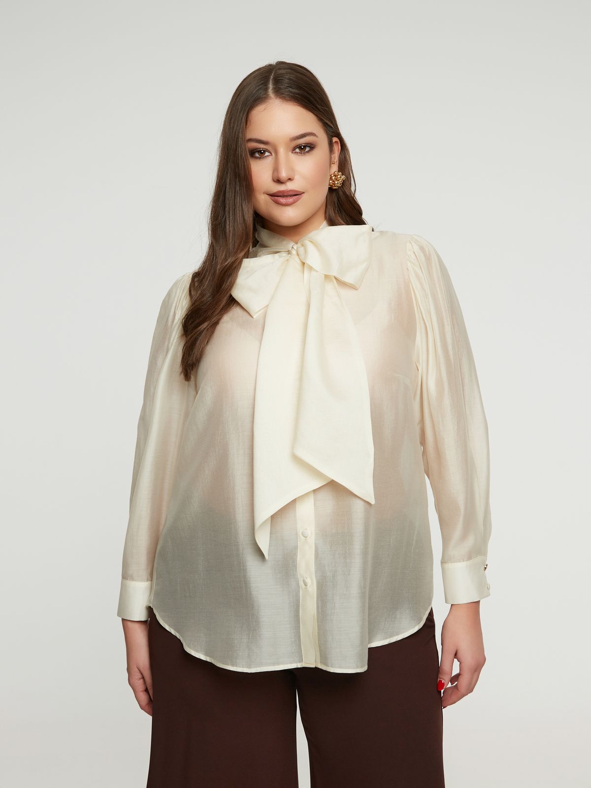 Een vrouw met lang bruin haar poseert voorover gericht tegen een witte achtergrond. Ze draagt de Crème hemd van Mat Fashion met een grote strik in de nek en een donkerbruine broek.