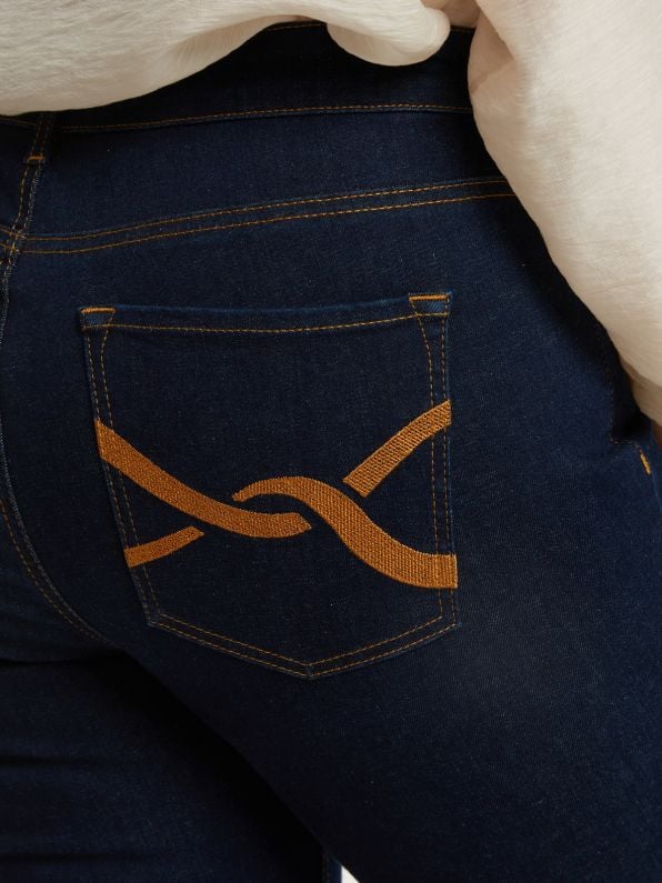 Close-up van de achterzak van donkerblauw Jeansbroek - Mat Fashion met oranje stiksel met lusjes. Een wit overhemd is gedeeltelijk zichtbaar aan de bovenkant.