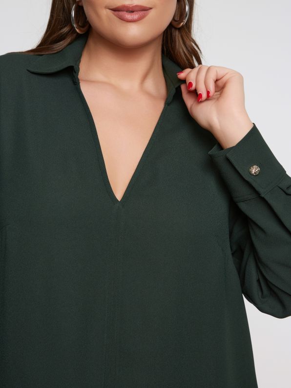 Een vrouw draagt het Groen kleed van Mat Fashion, met een donkergroene tint, V-hals en manchetten met knopen. Haar roodgelakte nagels bij haar kraag voegen een vleugje elegantie toe, waarbij haar gezicht gedeeltelijk zichtbaar is.